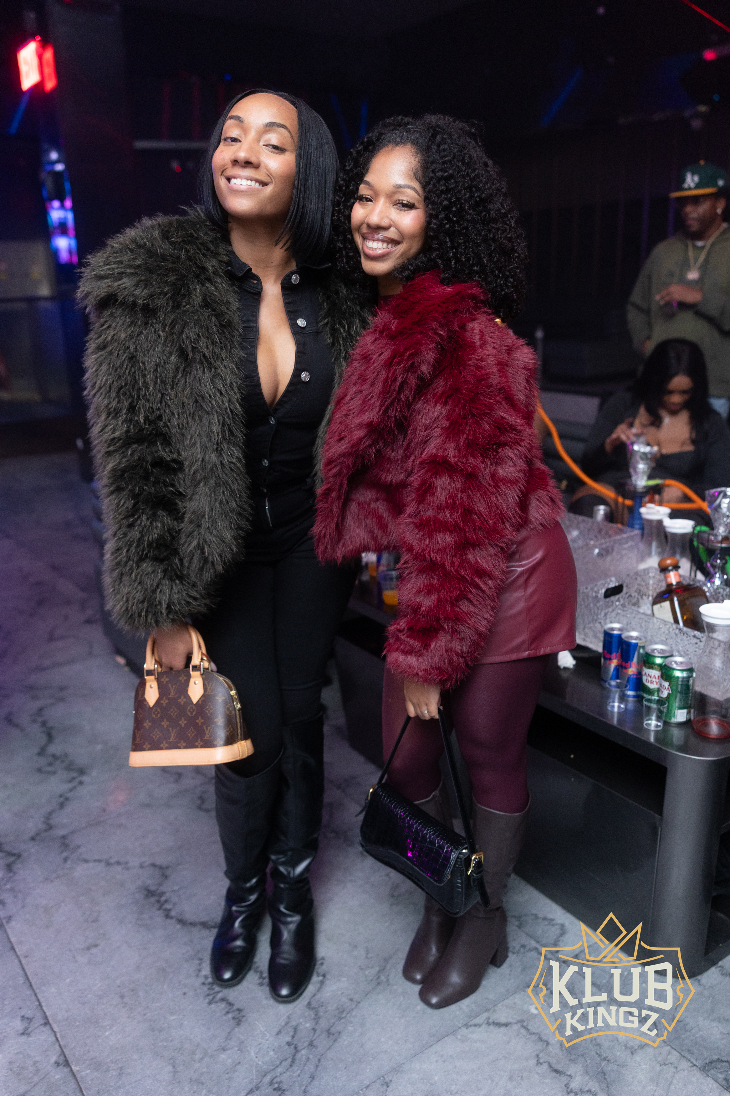 Two stylish women in faux fur jackets pose together at Klub Kingz nightclub event