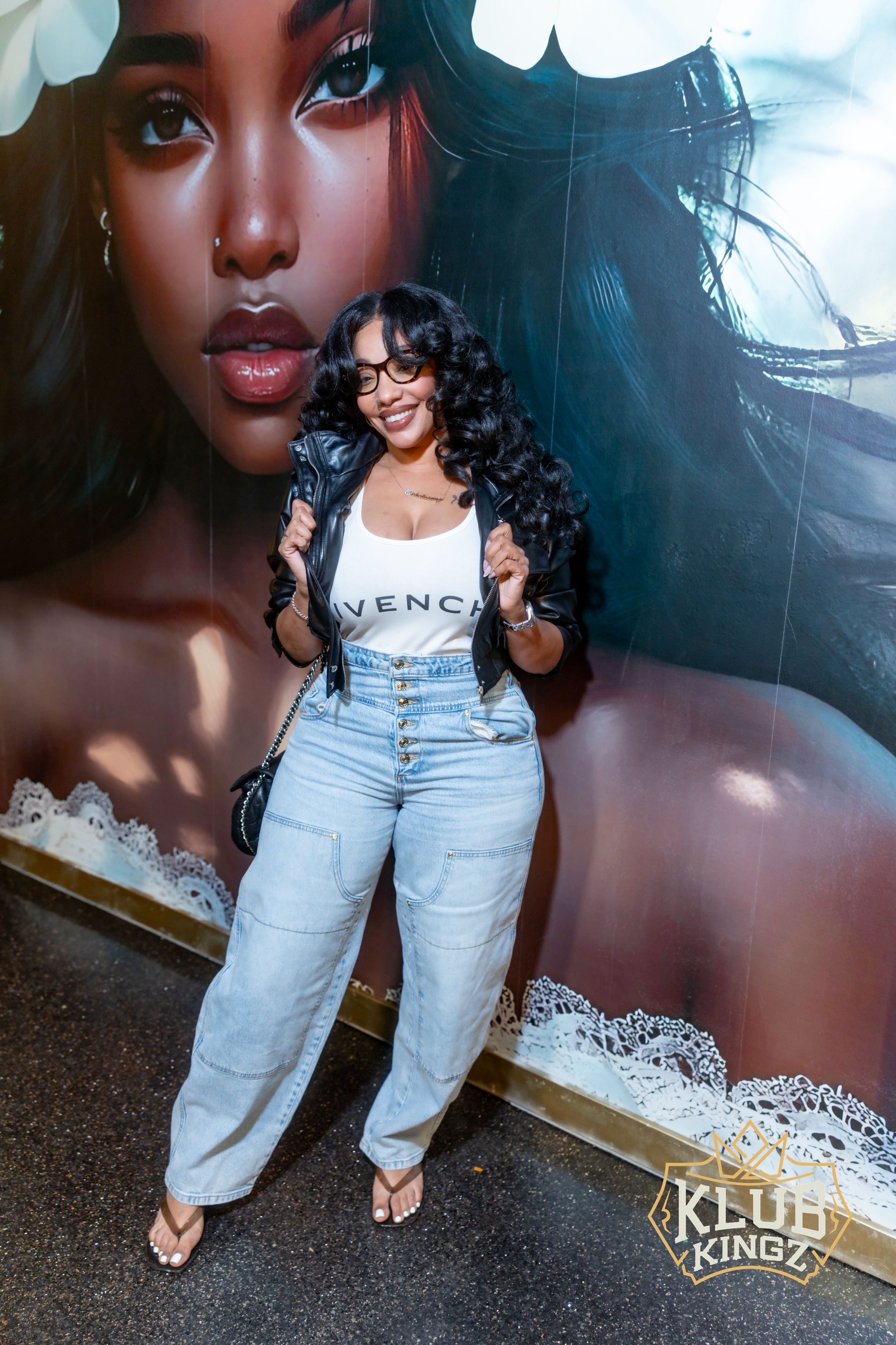 Smiling woman in leather jacket and jeans posing by mural at Klub Kingz nightlife event