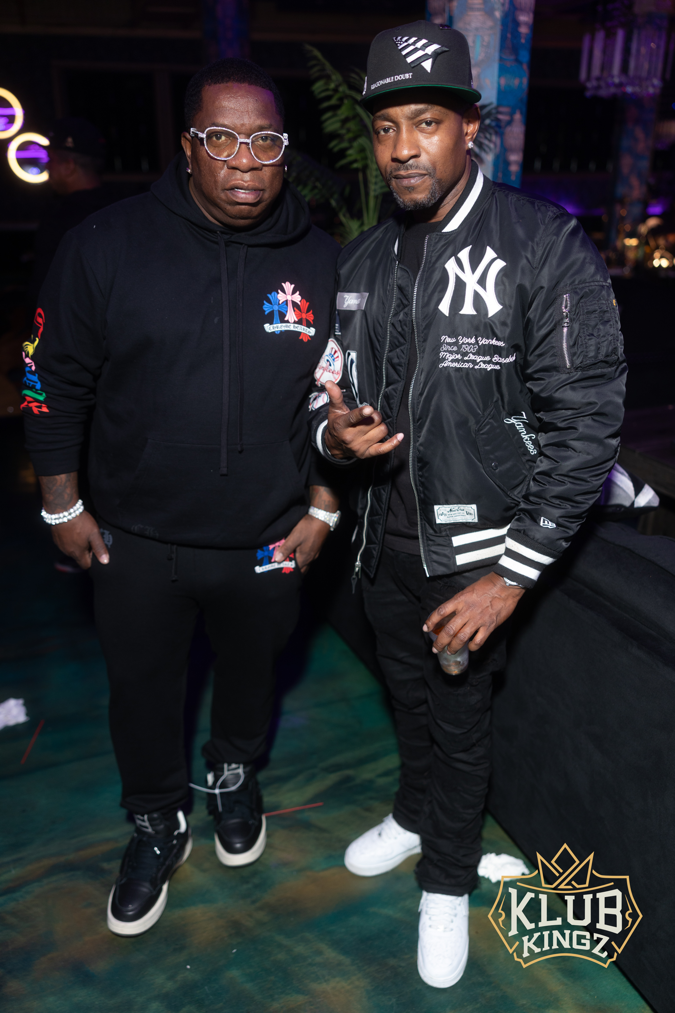 Two men pose together in streetwear and a Yankees jacket at a Klub Kingz nightlife event