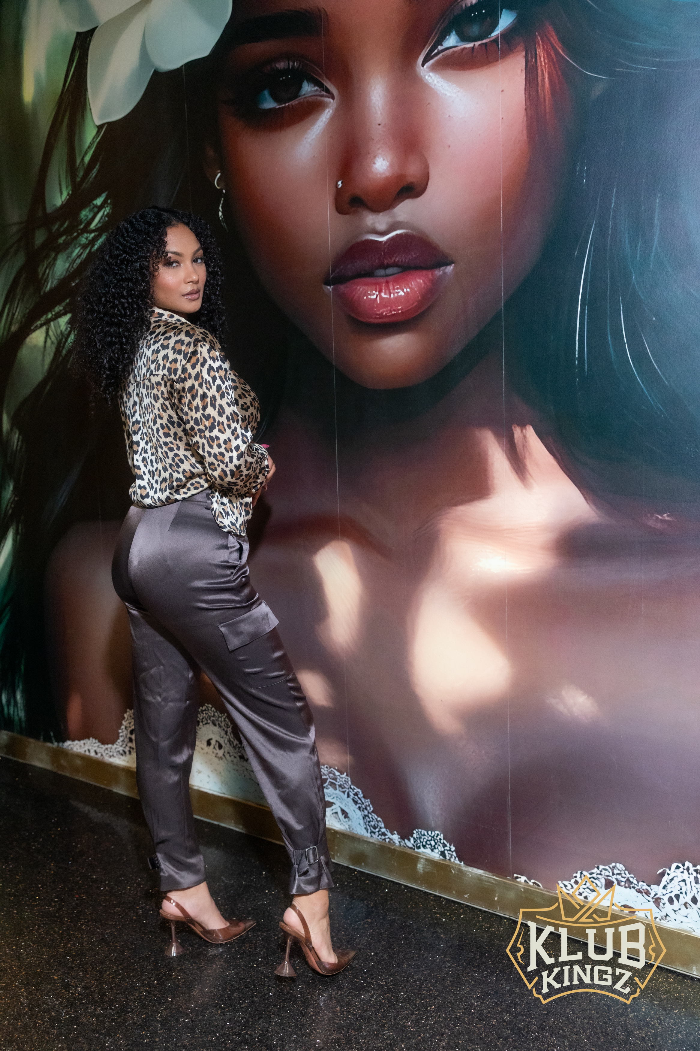 Woman in leopard-print top posing beside large portrait mural at Klub Kingz venue
