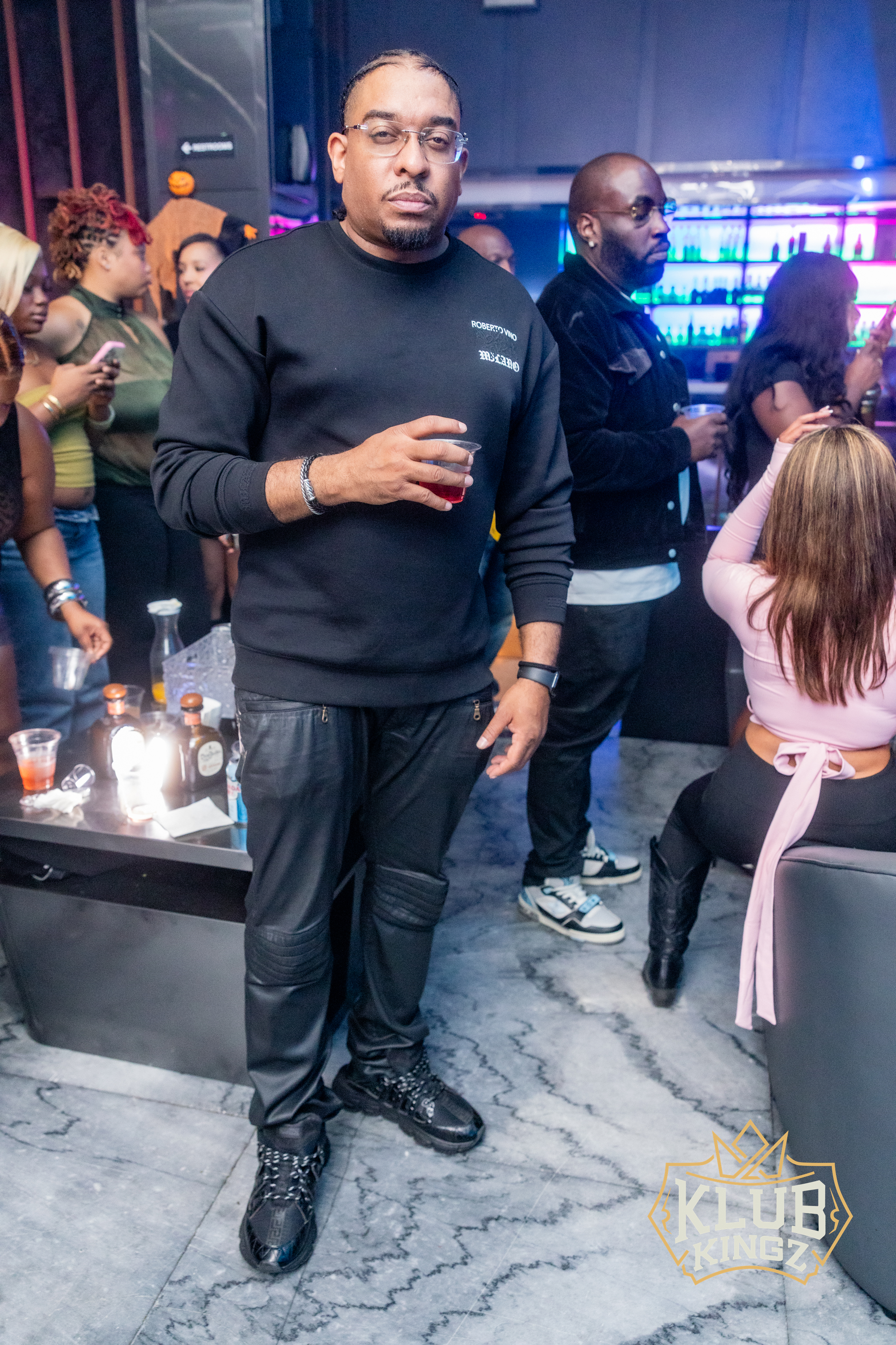 Man in black sweatshirt holding a drink at Klub Kingz nightclub with guests and bar in background